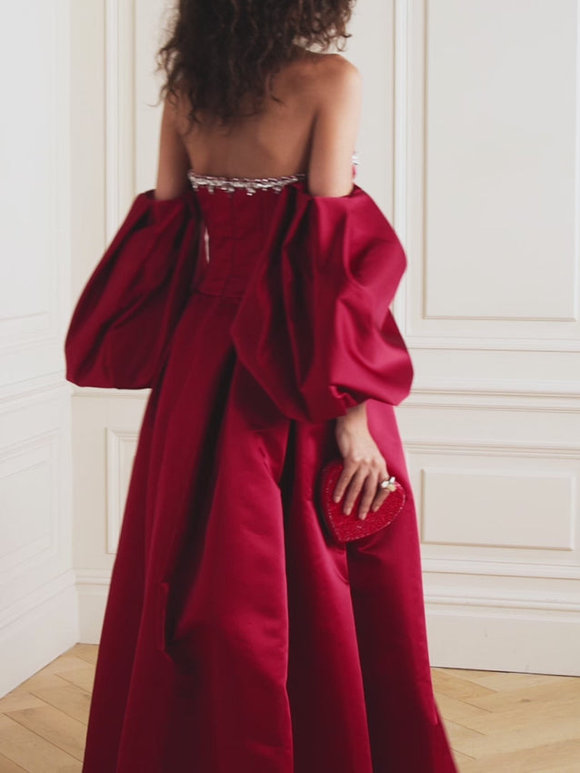 This stunning mikado gown transforms you into royalty. The off-the-shoulder silhouette, adorned with billowy sleeves, frames the neckline beautifully while the voluminous skirt creates an elegant sweep. Hand-scattered crystals catch the light across the rich red fabric, casting a mesmerizing glow with every step. Designed for those unforgettable moments when you want to feel absolutely magical.
