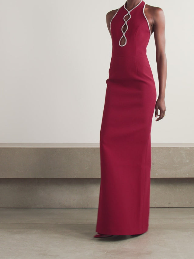 The Akari gown epitomizes modern elegance with its striking halter neckline and daring cutout design. Crafted from luxurious burgundy cady, this floor-length gown hugs your silhouette with a fitted waist that flatters and defines. Sparkling crystals trace the strategic cutouts, catching light with every movement. The piece reveals its true showstopper detail when you turn—an open back that exudes sophistication and allure. Perfect for red carpet moments and unforgettable evenings.