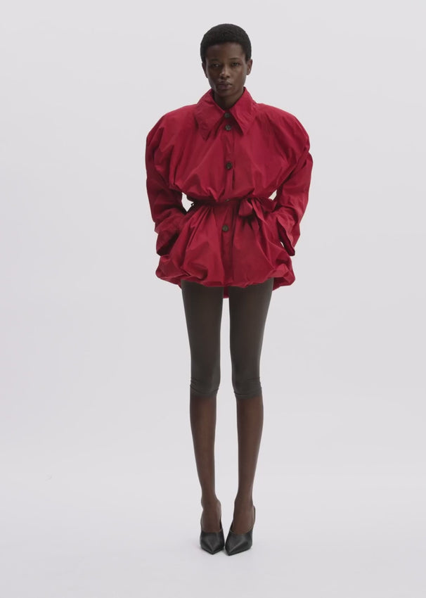 This striking jacket hits at the hip with a bold, sculptural design. The standout bubble hemline creates dramatic volume, while a stand collar and raglan sleeves define the silhouette. Practical front welt pockets and a belted waist—secured with a metal buckle through three loops—add both function and style. Inside, a luxurious silk lining provides comfort, and the front button closure completes the polished look.