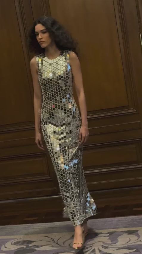 A show-stopping midi dress that commands attention with its reflective silver mirrored embroidery on delicate tulle. The jewel neckline frames the face beautifully while the form-fitting silhouette hugs your curves, with strategically placed mirrored motifs creating dimension and a sculpted look. Every movement catches light, ensuring you shimmer from every angle. Sleeveless and sophisticated—perfect for elevating your evening look.
